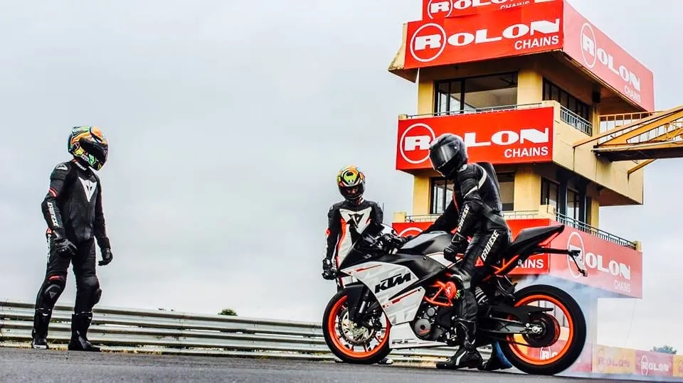 Three riders in leathers around an orange KTM in front of the timing tower